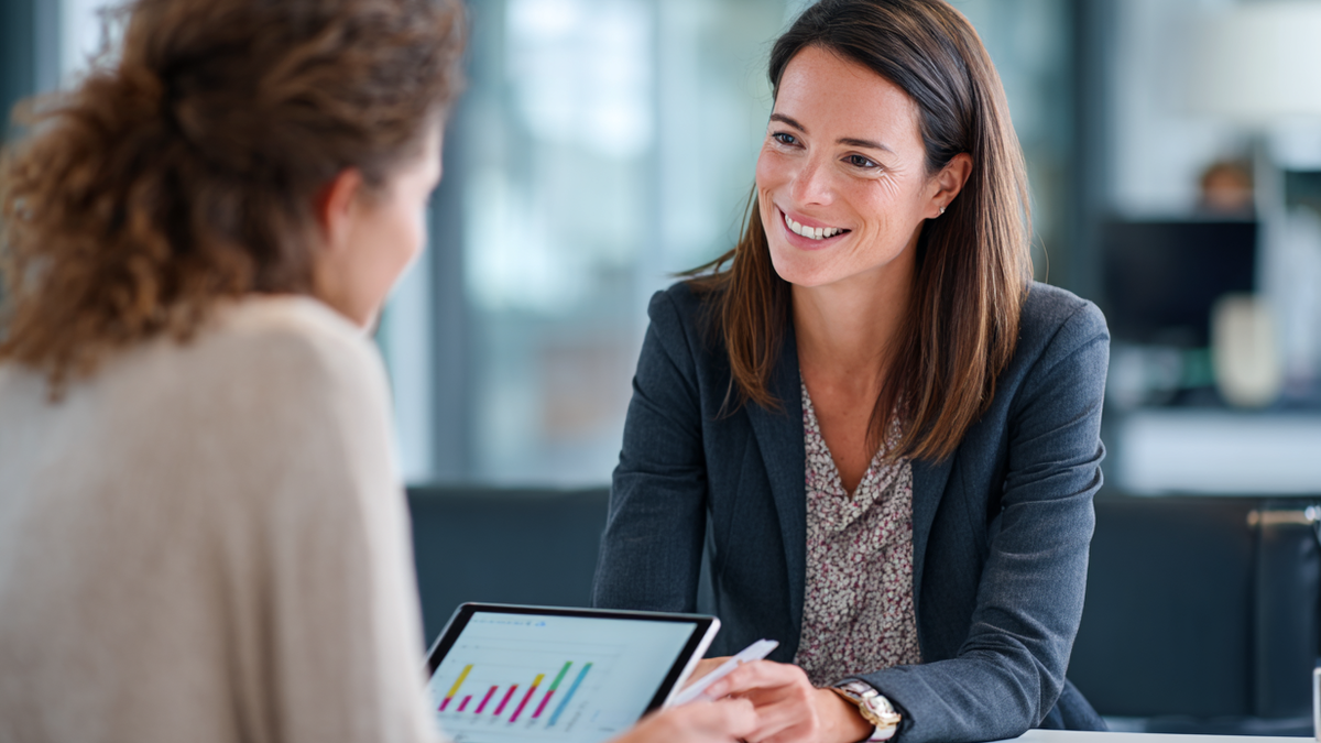 Junger Klient und Finanzberater besprechen lächelnd Inhalte auf Tablet an einem Schreibtisch im modernen Büro.