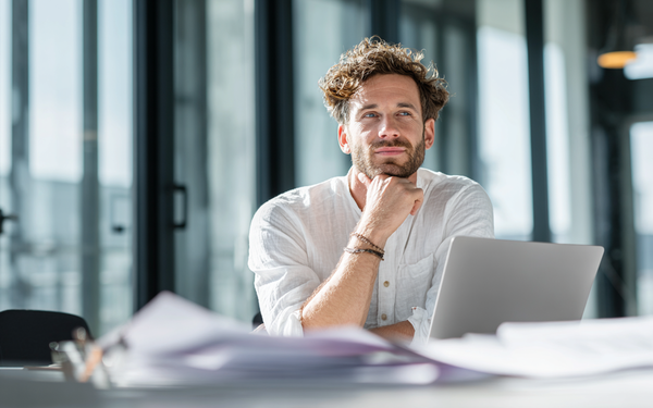 Mann mit Bart sitzt an einem Tisch vor seinem Laptop und wirkt nachdenklich aber zuversichtlich.