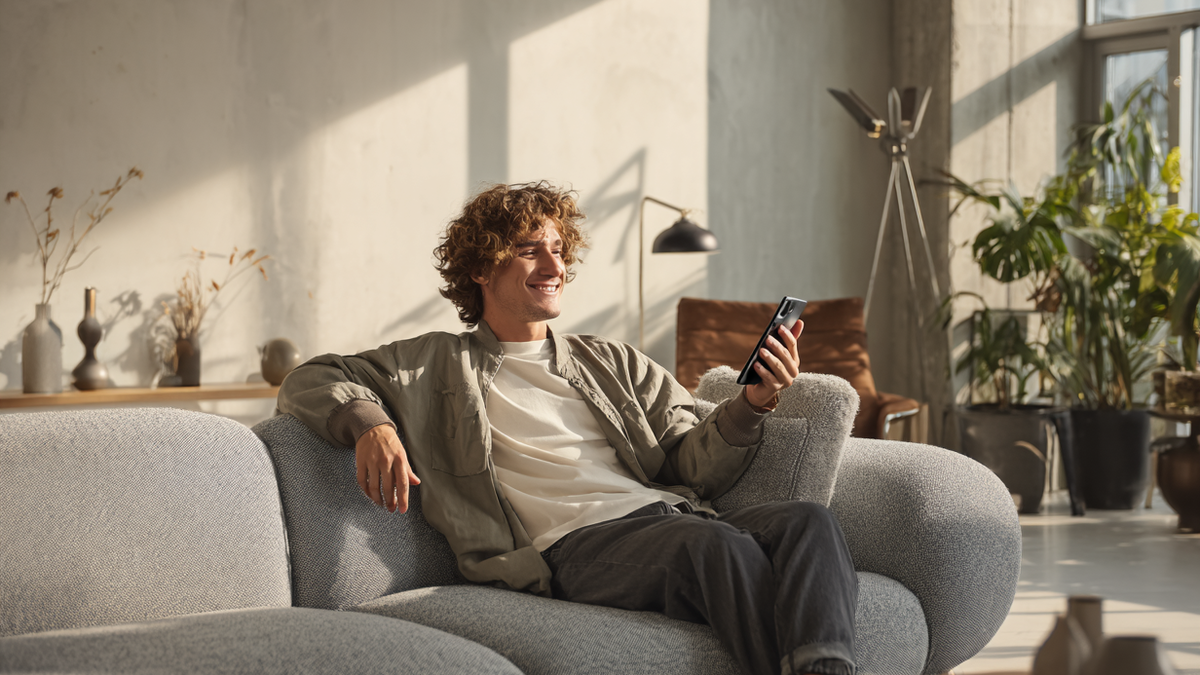 Mann sitzt auf Sofa im Wohnzimmer und überprüft glücklich Tagesgeldzinsen auf ihrem Smartphone.