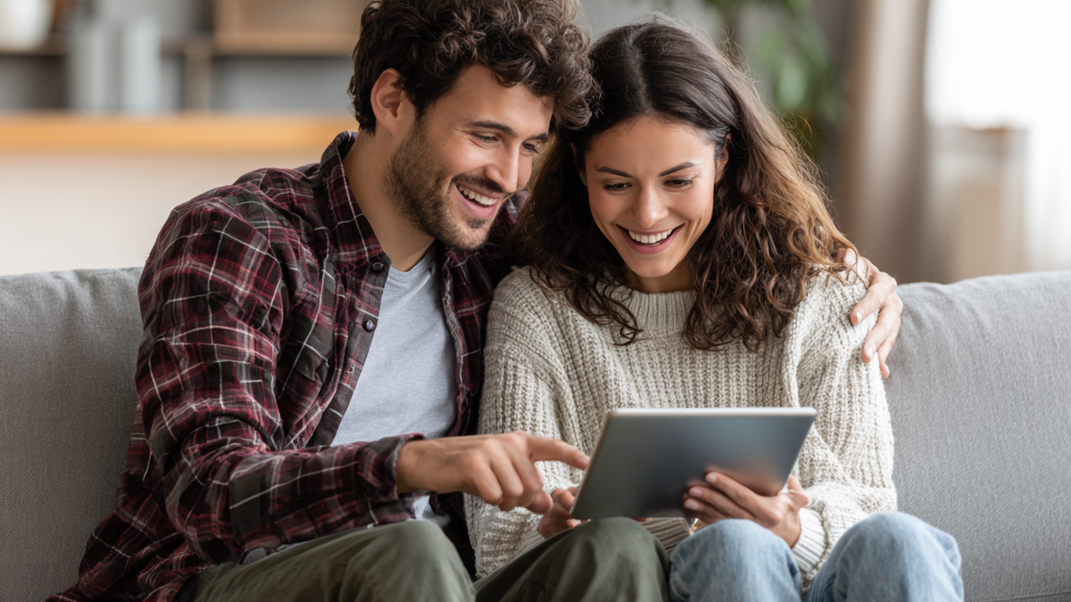 Paar sitzt gemütlich auf Sofa und blickt lächelnd gemeinsam auf Tablet, um sein Gemeinschaftskonto zu verwalten