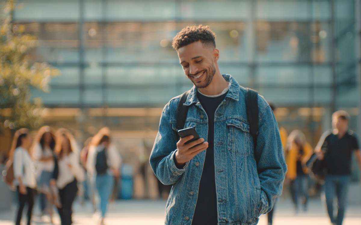 Junger Student steht vor einem Unigebäude mit einem Smartphone in der Hand
