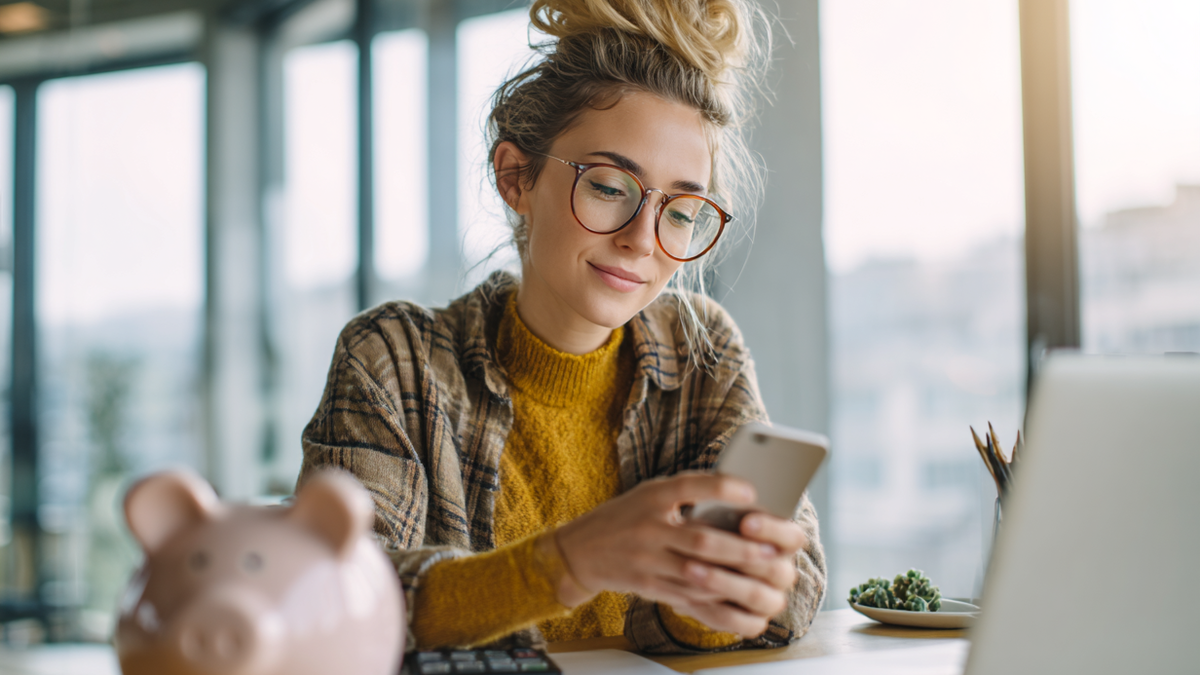 Junge Frau sitzt an einem Schreibtisch mit einem Sparschwein und schaut auf ein Handy 