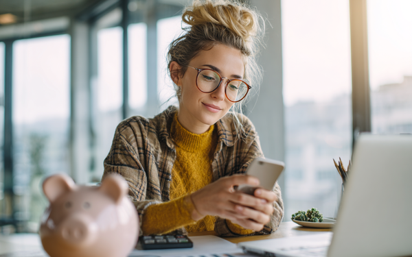Junge Frau sitzt an einem Schreibtisch mit einem Sparschwein und schaut auf ein Handy 