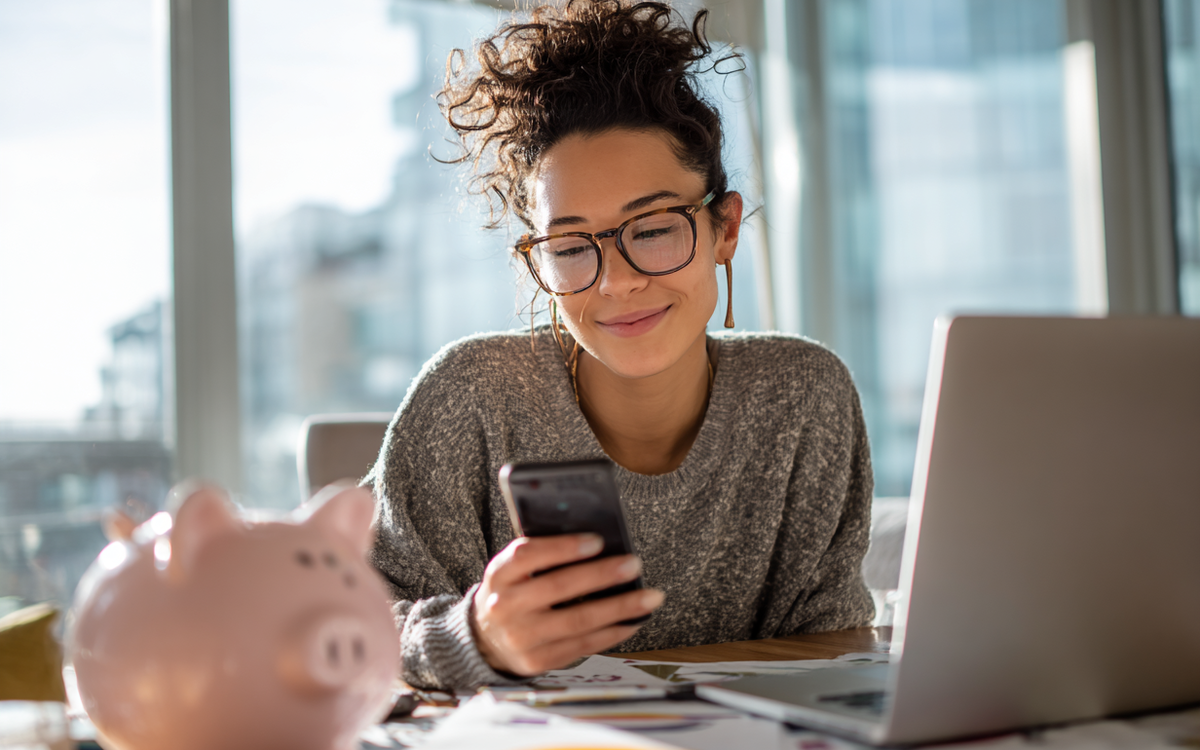Junge Frau sitzt vor einem Laptop an einem Schreibtisch mit Sparschwein, hält ein Handy in der Hand