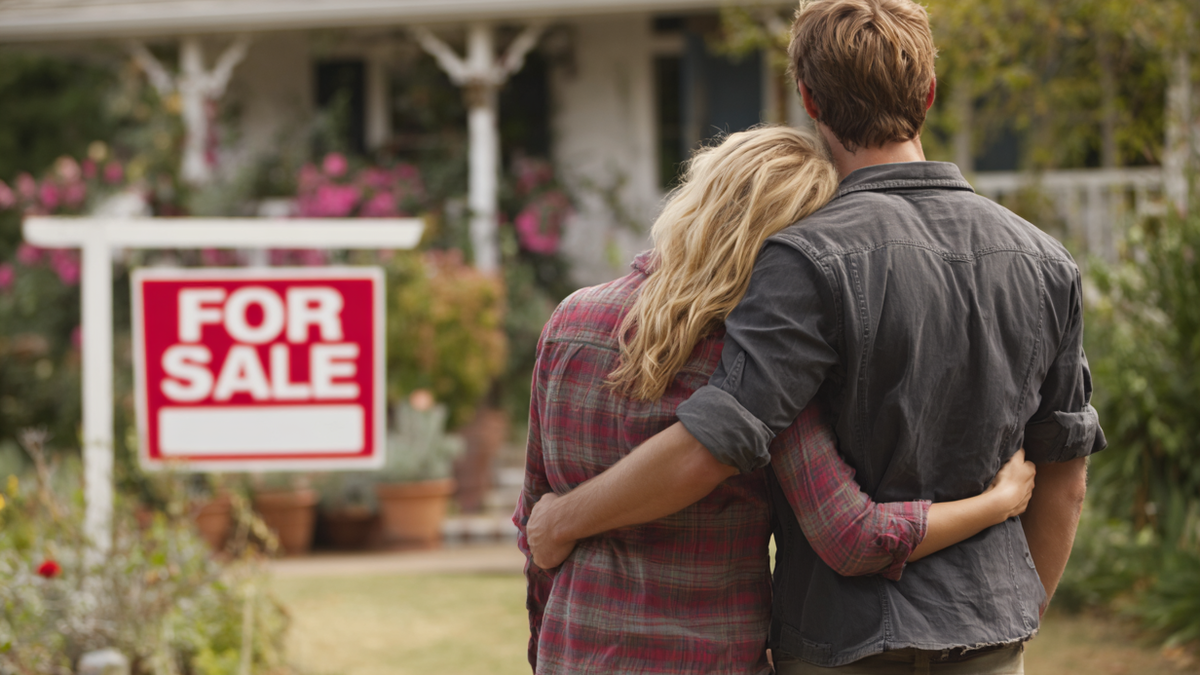 Ein junges Paar steht vor einem Haus, an dem deutlich "For Sale" steht