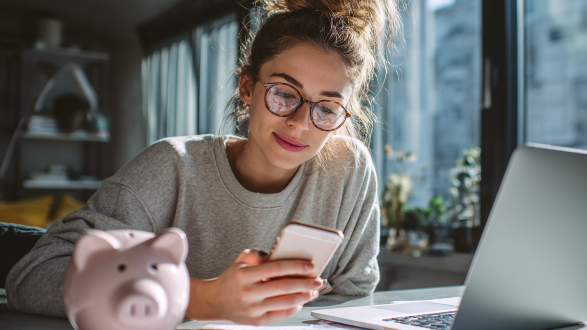 Junge Frau sitzt an einem Schreibtisch mit einem Sparschwein und schaut auf ein Handy 