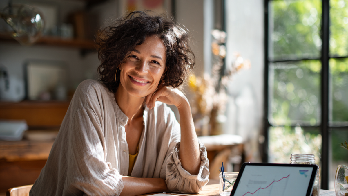 Frau sitzt lächelnd an einem Tisch, auf dem ein Tablet steht was einen steigenden Graphen zeigt