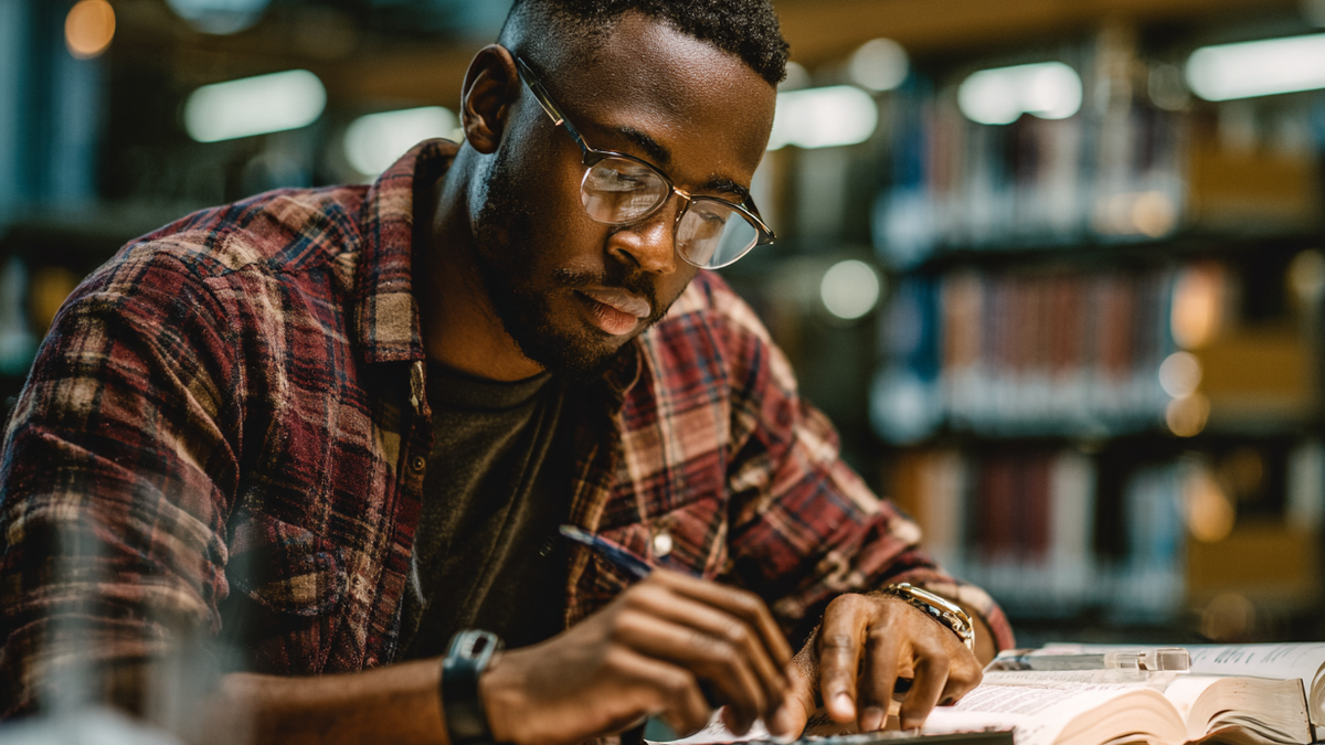 Junger, farbiger Mann sitzt an einem Schreibtisch in einem Bibliothek und tippt auf einem Taschenrechner